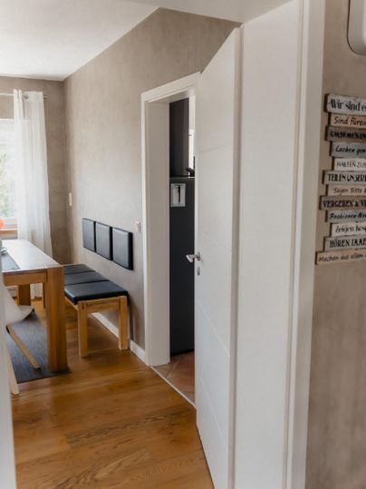 A bright hallway leading to an open space with a table and chairs. A few pictures hang on the wall, and there is an open door leading to another room.