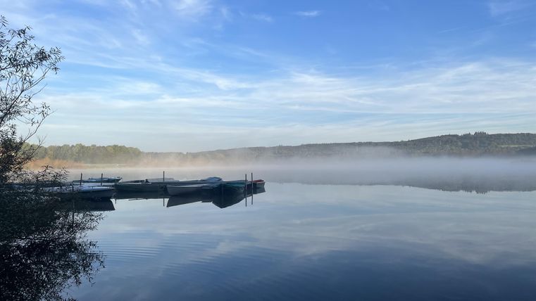 Ein ruhiger See mit Booten und einer leichten Nebeldecke. Der Himmel ist klar und blau, was eine friedliche Stimmung vermittelt.