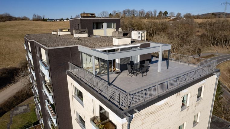 Eine moderne Wohnanlage mit einem großzügigen Balkon. Im Hintergrund sind sanfte Hügel und ein klarer Himmel zu sehen.