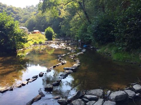 Ein ruhiger Fluss verläuft durch einen bewaldeten Bereich, mit flachen Steinen, die im Wasser liegen. Im Hintergrund sind Bäume und einige Zelte sichtbar.