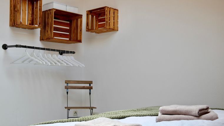 Ein schlichtes Zimmer mit einem Bett und mehreren Handtüchern. An der Wand hängen Holzregale und eine Kleiderstange mit Kleiderbügeln.