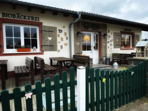 Ein einladendes Biobäckerei-Gebäude mit Holzverzierungen und einem grünen Zaun. Vor dem Eingang stehen Tische und Stühle zum Verweilen.