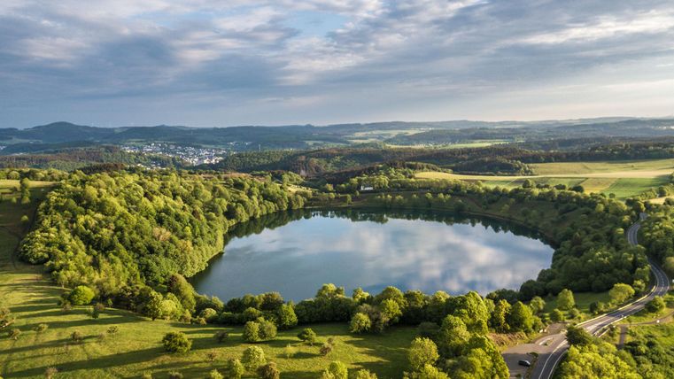 Ein malerischer See umgeben von üppigem Grün und sanften Hügeln. Der Himmel ist wechselhaft mit Wolken und einem klaren Blau.