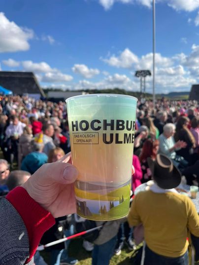 Eine Hand hält ein Getränk in einem Becher mit der Aufschrift "HOCHBAU ULM". Im Hintergrund ist eine große Menschenmenge unter einem blauen Himmel zu sehen.