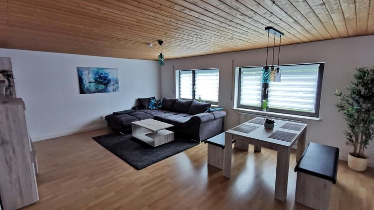 Ein modernes Wohnzimmer mit einem gemütlichen Sofa und einem Esstisch. Große Fenster lassen viel Licht herein und es gibt eine Pflanze in der Ecke.