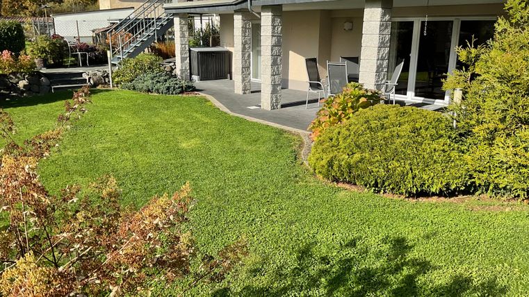 Ein gepflegter Garten mit grünem Rasen und bunten Sträuchern umgibt ein modernes Haus. Auf der Terrasse stehen Gartenmöbel, ideal für entspannte Stunden im Freien.