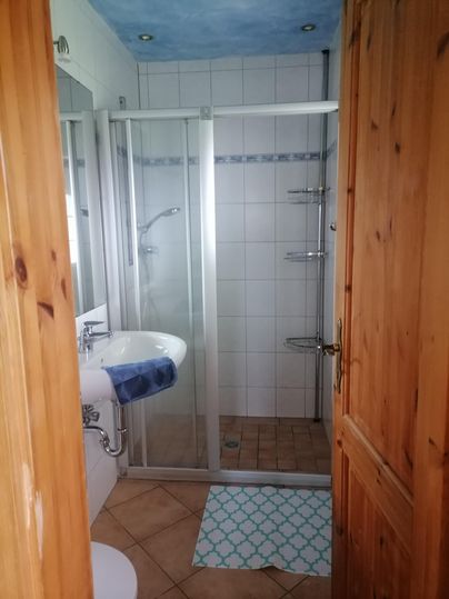 Ein helles Badezimmer mit einer Dusche, einem Waschbecken und Fliesenboden. Die Wände sind gefliest, und es gibt eine Holz-Tür.