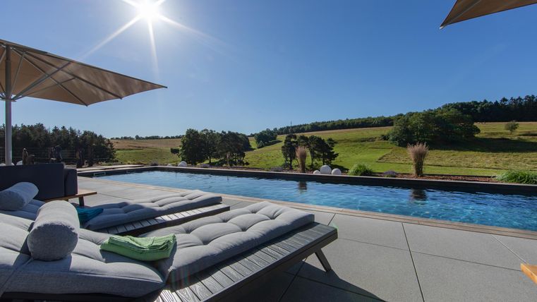 Ein wunderschöner Swimmingpool mit Liegen und Sonnenschirmen. Im Hintergrund erstreckt sich eine grüne Landschaft unter klarem blauen Himmel.