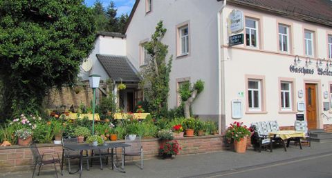 Ein charmantes Gasthaus mit blühendem Garten und gemütlichen Sitzplätzen im Freien. Die Fassade ist freundlich und einladend gestaltet.
