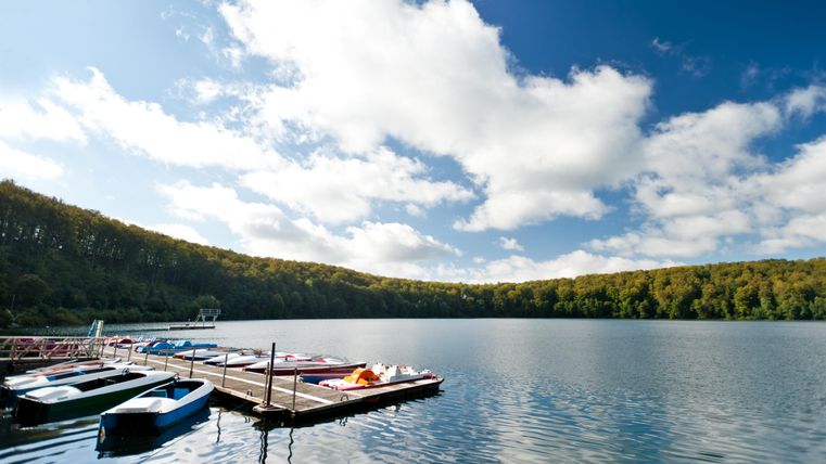 Ein ruhiger See mit einem Steg und mehreren Booten. Im Hintergrund sind grüne Bäume und ein klarer blauer Himmel zu sehen.