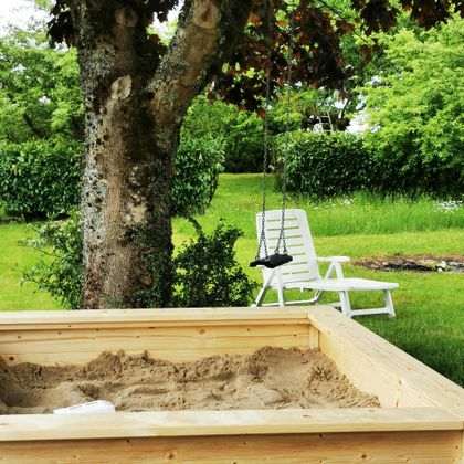Ein Sandkasten aus Holz und eine Schaukel stehen im Garten unter einem großen Baum. Im Hintergrund sind grüne Sträucher und eine Liege zu sehen.