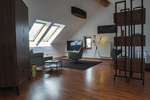 Ein modernes Wohnzimmer mit schrägen Fenstern und Holzboden. Die Einrichtung umfasst einen gemütlichen Sessel, einen Tisch und einen Fernseher.