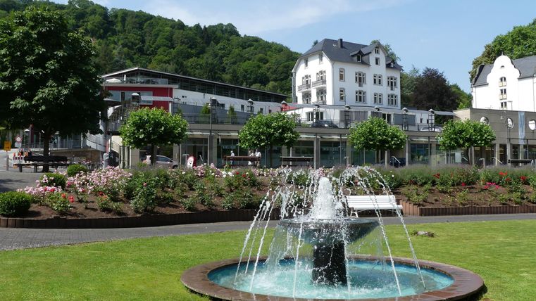 Ein schöner Springbrunnen in einem gepflegten Park. Im Hintergrund sind Gebäude und grüne Bäume zu sehen.