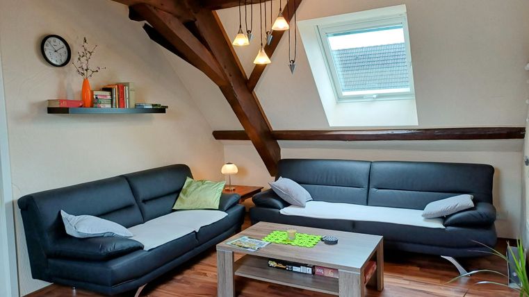 Ein gemütliches Wohnzimmer mit zwei schwarzen Sofas und einem Holztisch. Große Fenster lassen viel Licht herein und die Deckenbalken verleihen dem Raum einen besonderen Charme.