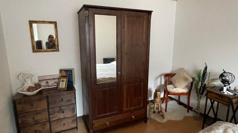 Ein gemütliches Schlafzimmer mit einem klassischen Kleiderschrank und einem Nachttisch. Auf der Kommode liegen persönliche Gegenstände und eine kleine Pflanze steht neben dem Stuhl.