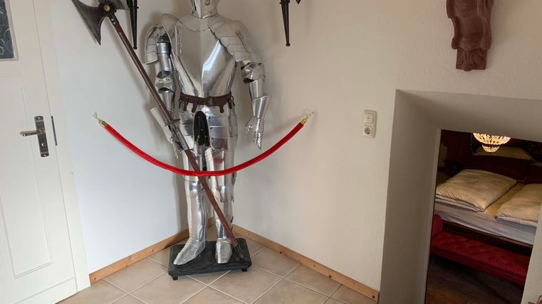 Ein Ritter in glänzender Rüstung steht an der Wand neben einem roten Absperrband. Im Hintergrund ist ein geschmackvoll eingerichteter Raum mit einem Bett sichtbar.