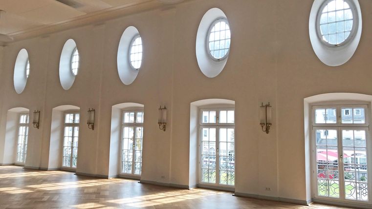 Ein heller Raum mit ovalen Fenstern und großen Türen. Tageslicht fällt auf den eleganten Holzfußboden.