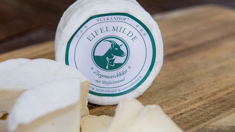 Ein runder Käse mit der Aufschrift "Eifelmilde" auf einem Holzbrett. Er ist in Stücke geschnitten und hat eine cremige Textur.