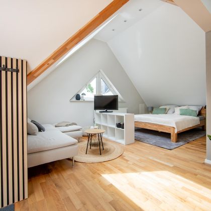 Ein modernes Zimmer mit einem gemütlichen Sofa und einem großen Fenster. Der Boden ist aus Holz und es gibt eine entspannte Atmosphäre.