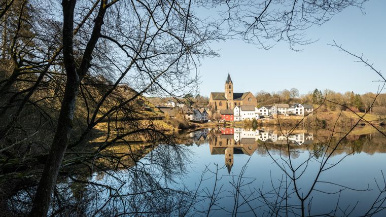 A peaceful landscape with a clear lake that reflects the church and the surrounding buildings. Trees and a clear sky can be seen in the background.