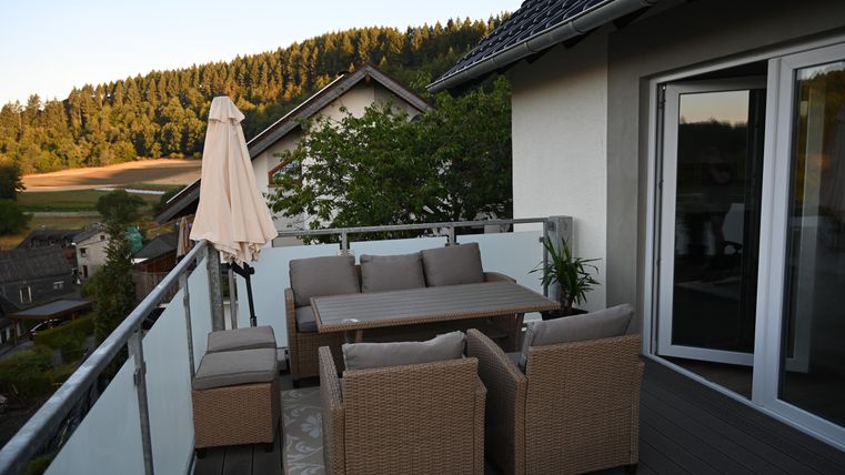 Eine gemütliche Terrasse mit einem Esstisch und Stühlen aus Rattan. Im Hintergrund sind Bäume und Hügel zu sehen, die eine ruhige Landschaft bilden.