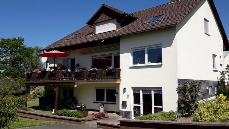 Ein schönes Haus mit einem Balkon und rotem Sonnenschirm. Es ist von einem gepflegten Garten umgeben und der Himmel ist blau.