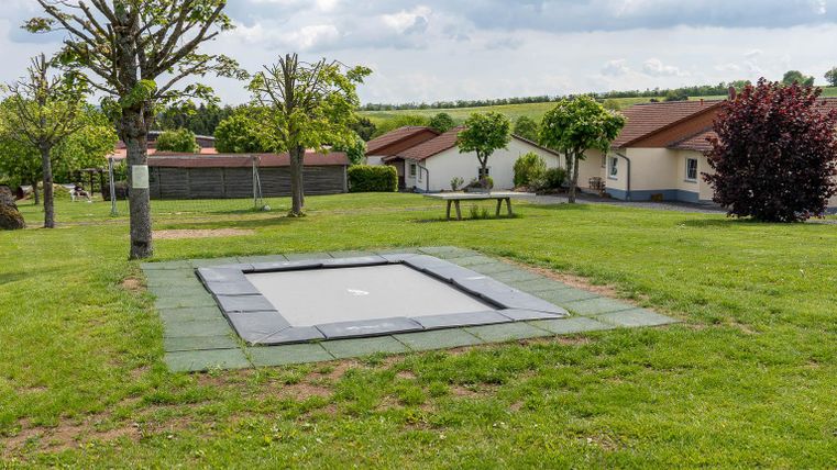 Ein Trampolin auf einer grünen Wiese, umgeben von Bäumen und Häusern. Der Himmel ist bewölkt, und die Umgebung wirkt ruhig und einladend.