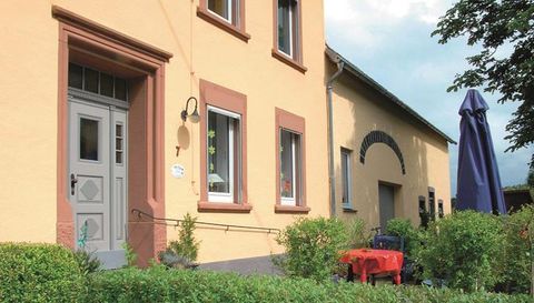 Ein charmantes Haus mit hellgelber Fassade und einer einladenden Eingangstür. Im Vordergrund sind grüne Büsche und ein roter Tisch zu sehen.