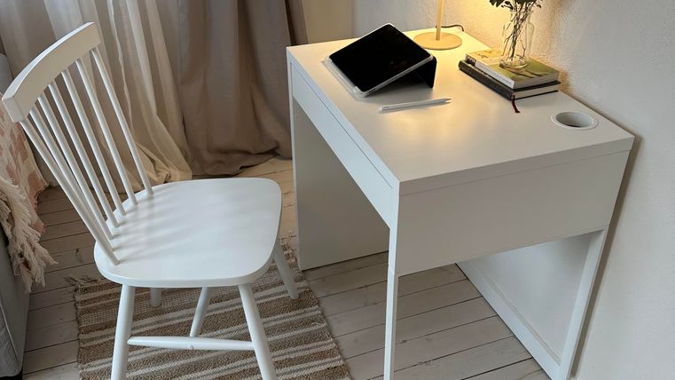 Ein einfacher Schreibtisch mit einer Lampe, einem Tablet und einer kleinen Vase mit Blumen. Neben dem Schreibtisch steht ein Holzstuhl und auf dem Boden liegt ein Teppich.