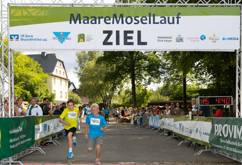 Zwei Jungen laufen mit Begeisterung über die Ziellinie eines Wettlaufs. Im Hintergrund sind Zuschauer und eine grüne Zielbanner zu sehen.