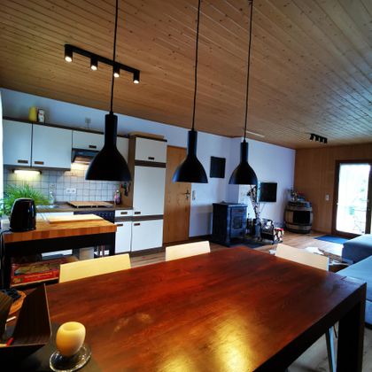 Eine moderne Küche mit einem großen Esstisch aus Holz und schwarzer Beleuchtung. Der Raum ist hell und freundlich, mit einem Zugang zu einem Außenbereich.