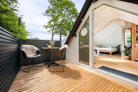 Eine moderne Terrasse mit Holzbelag und zwei Stühlen. Im Hintergrund ist ein helles Zimmer mit einem Bett und Pflanzen sichtbar.