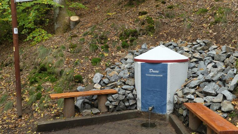 Ein kleiner Wasserbrunnen in der Natur, umgeben von Steinen und Bänken. Die Umgebung ist grün und zeigt viel Erd- und Pflanzenboden.