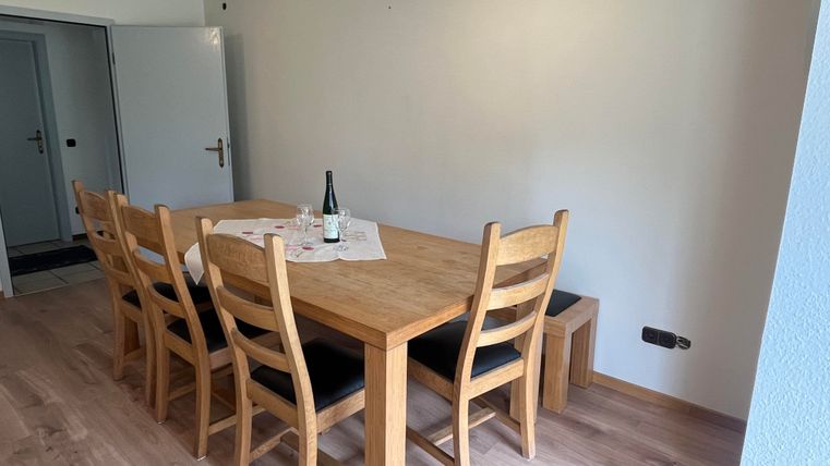 Ein helles Esszimmer mit einem großen Holztisch und sechs Stühlen. Auf dem Tisch steht eine Flasche Wein und ein Tischläufer.