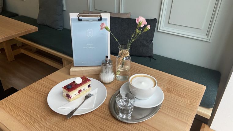 Ein Tisch in einem Café mit einem Stück Kuchen und einer Tasse Kaffee. Eine kleine Vase mit einer Blume steht neben dem Dessert.
