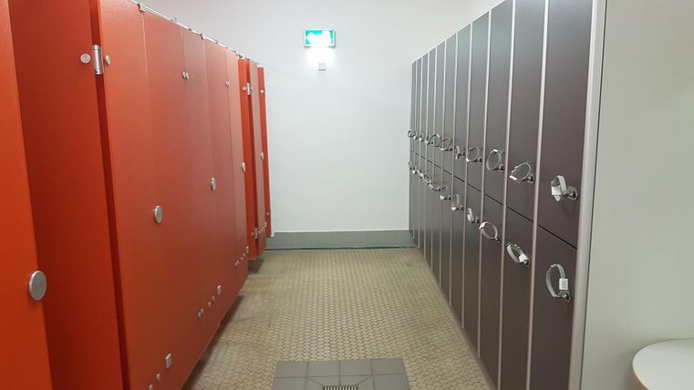 Ein Umkleideraum mit orangefarbenen und grauen Schließfächern. Der Raum hat eine helle Beleuchtung und einen gefliesten Boden.