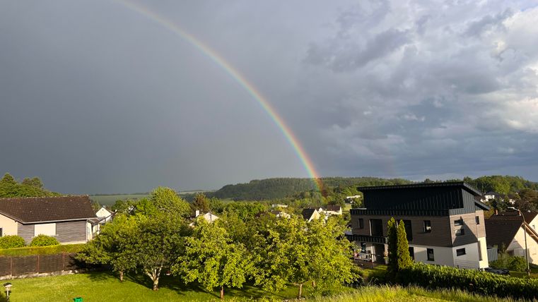 Ein Regenbogen erscheint über einem ruhigen Landschaftsbild mit Häusern und grünen Bäumen. Der Himmel ist bewölkt, aber die Farben des Regenbogens leuchten deutlich hervor.