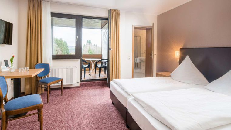Ein modernes Hotelzimmer mit zwei Einzelbetten, einem Tisch und Stühlen. Große Fenster bieten einen Blick auf die Außenanlage.