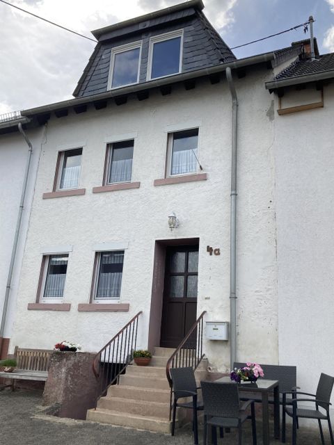 Ein zweistöckiges weißes Haus mit einem Satteldach. Vor dem Eingang stehen einige Gartenmöbel für die Terrasse.