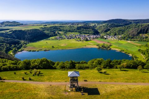 Eine malerische Landschaft mit einem klaren See und umgeben von grünen Wiesen. Im Hintergrund sind sanfte Hügel und ein kleines Dorf zu sehen.
