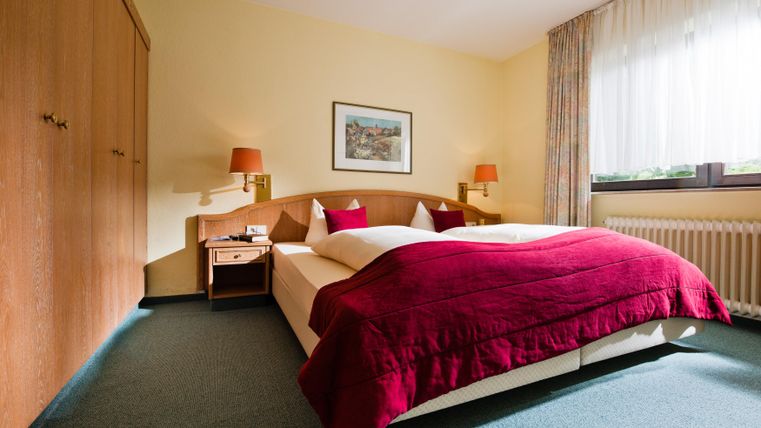 Ein modernes Schlafzimmer mit einem großen Bett und roten Bettdecken. Die Wände sind in einem sanften Gelbton gestrichen und es gibt einen Kleiderschrank sowie einen Nachttisch.
