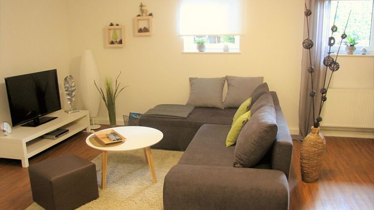 Ein gemütliches Wohnzimmer mit einem grauen Sofa und einem runden Tisch. Der Raum hat einen Fernseher und helle Fenster mit Pflanzen.