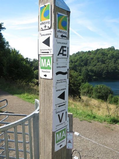 Ein Wegweiser mit verschiedenen Schildern auf einem Wanderweg. Im Hintergrund ist ein schöner See und grüne Bäume zu sehen.