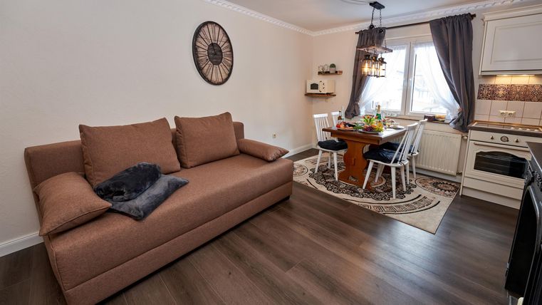 Ein gemütliches Wohnzimmer mit einem braunen Sofa und einem Esstisch. Die Fenster lassen viel Tageslicht herein und die Einrichtung ist modern und einladend.
