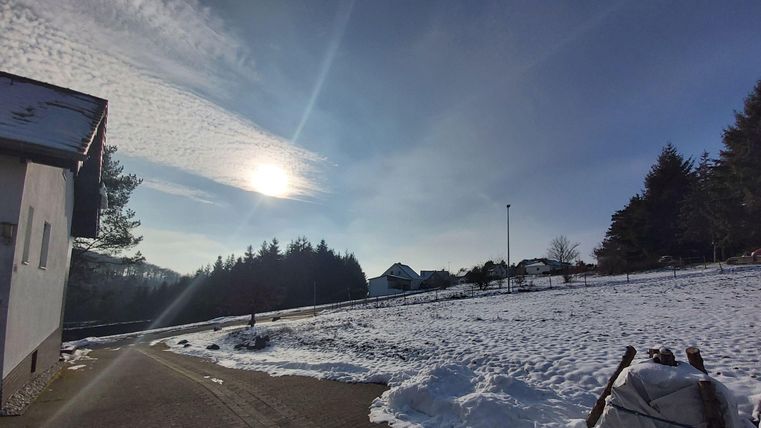 Eine schneebedeckte Landschaft unter einem klaren Himmel. Die Sonne strahlt über die Bäume und Wiesen.