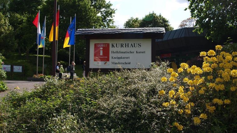 Ein Eingangsbereich mit dem Schild „Kurhaus“ und bunten Flaggen. Im Vordergrund blühen gelbe Blumen.