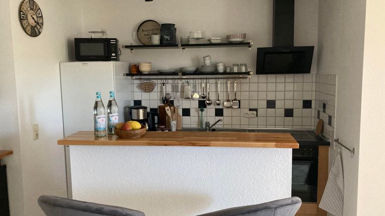 Eine moderne Küche mit einem Holzbar und zwei Barhockern. Auf der Arbeitsfläche stehen Wasserflaschen und Obst, während die Regale gut sortiert sind.