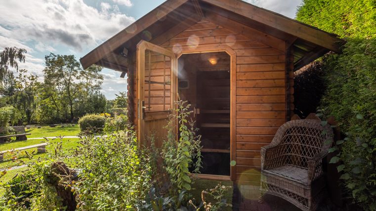 Eine kleine Holzsauna mit offener Tür steht in einem grünen Garten. Neben der Sauna befindet sich eine gemütliche Rattanbank.