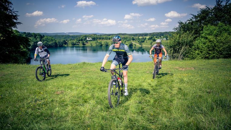 Drei Mountainbiker fahren entlang eines grünen Hügels mit Blick auf einen See. Der Himmel ist blau und mit einigen Wolken durchzogen.