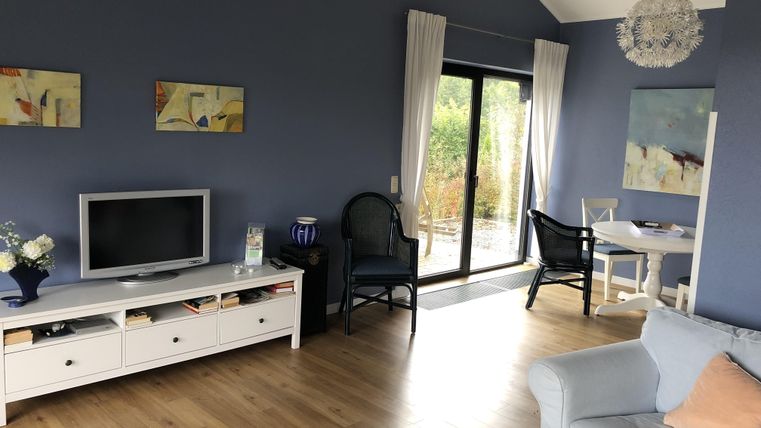 Ein modernes Wohnzimmer mit blauen Wänden und Holzfußboden. Es gibt ein TV-Möbel, einen gemütlichen Sessel und einen Esstisch mit Stühlen.